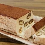 A rectangular slice of tiramisu semifreddo is displayed on a white dish. The dessert features creamy layers interspersed with small pieces of sponge cake. The top is dusted with cocoa powder, and two slices have been cut from the main loaf, showing its interior.