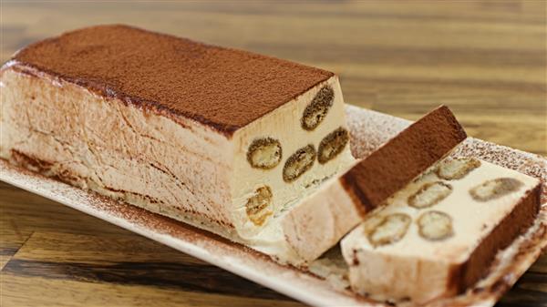 A rectangular slice of tiramisu semifreddo is displayed on a white dish. The dessert features creamy layers interspersed with small pieces of sponge cake. The top is dusted with cocoa powder, and two slices have been cut from the main loaf, showing its interior.