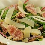 A plate of fresh salad containing sliced figs, arugula, and shaved Parmesan cheese. The ingredients are arranged in a decorative cream-colored bowl with scalloped edges and floral details. The focus is on the vibrant ingredients and textures of the salad.