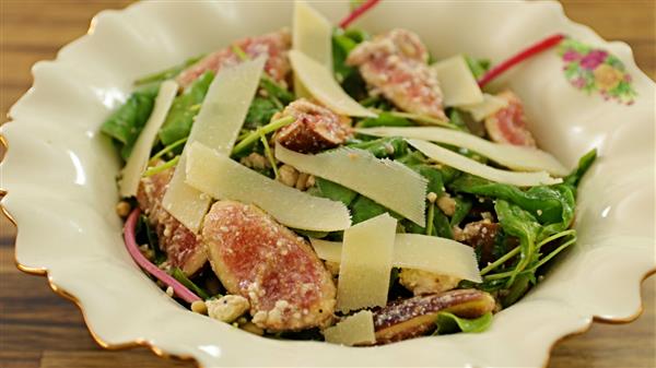 A plate of fresh salad containing sliced figs, arugula, and shaved Parmesan cheese. The ingredients are arranged in a decorative cream-colored bowl with scalloped edges and floral details. The focus is on the vibrant ingredients and textures of the salad.