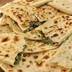 A silver tray holding several pieces of flatbread stuffed with green herbs and cheese. The flatbreads are golden-brown with char marks, and some pieces are stacked while others are partially folded, revealing the filling inside. The tray is on a wooden surface.