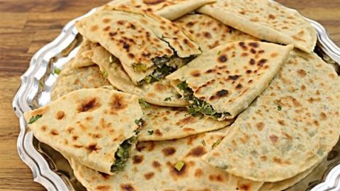 A silver tray holding several pieces of flatbread stuffed with green herbs and cheese. The flatbreads are golden-brown with char marks, and some pieces are stacked while others are partially folded, revealing the filling inside. The tray is on a wooden surface.