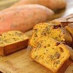 A loaf of sweet potato bread sliced on a wooden cutting board. Two slices are displayed in the foreground, showing a moist, orange interior studded with walnuts. Whole sweet potatoes are in the background on the wooden surface.