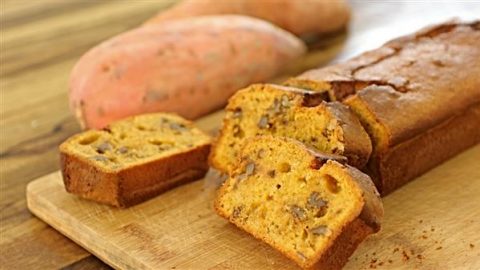 A loaf of sweet potato bread sliced on a wooden cutting board. Two slices are displayed in the foreground, showing a moist, orange interior studded with walnuts. Whole sweet potatoes are in the background on the wooden surface.