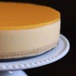 A smooth, round cheesecake with a glossy, golden yellow topping sits on a white, decorative cake stand. The cake has a creamy beige layer and a thin, crumbly crust at the bottom. The background is dark, making the cheesecake stand out.