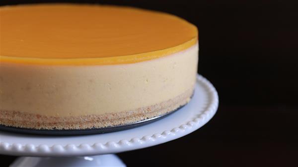 A smooth, round cheesecake with a glossy, golden yellow topping sits on a white, decorative cake stand. The cake has a creamy beige layer and a thin, crumbly crust at the bottom. The background is dark, making the cheesecake stand out.