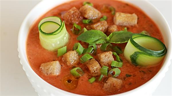 A bowl of gazpacho garnished with croutons, sliced green onions, fresh basil leaves, and cucumber ribbons. The cold soup has a vibrant red color and is presented in a white bowl.