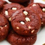 A plate of freshly baked red velvet cookies with white chocolate chips. The cookies have a deep red color and a cracked surface, showcasing some visible white chocolate chips. The plate is set on a wooden surface.