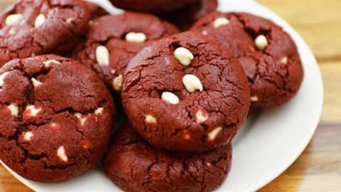 A plate of freshly baked red velvet cookies with white chocolate chips. The cookies have a deep red color and a cracked surface, showcasing some visible white chocolate chips. The plate is set on a wooden surface.