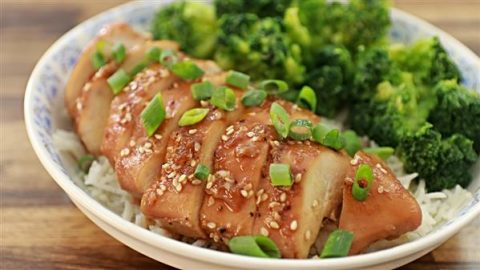 A bowl of sliced teriyaki chicken garnished with sesame seeds and green onions, served over a bed of white rice. Fresh, steamed broccoli florets are placed alongside the chicken. The dish is plated in a white bowl on a wooden surface.