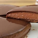 A rich, chocolate cheesecake with a glossy chocolate ganache topping is being served. A slice is lifted off the plate, revealing the smooth and creamy texture of the cheesecake. A jar of Nutella is visible in the background.