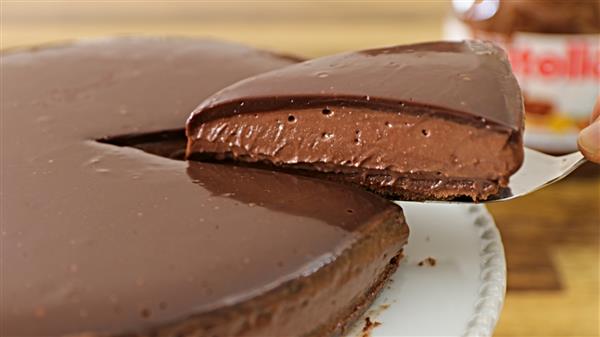 A rich, chocolate cheesecake with a glossy chocolate ganache topping is being served. A slice is lifted off the plate, revealing the smooth and creamy texture of the cheesecake. A jar of Nutella is visible in the background.