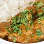 A close-up of a plate of creamy chicken curry topped with fresh cilantro, served with a side of white basmati rice. The rich, orange-brown sauce is visible with chunks of chicken, contrasting with the fluffy, plain white rice.