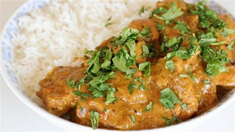 A close-up of a plate of creamy chicken curry topped with fresh cilantro, served with a side of white basmati rice. The rich, orange-brown sauce is visible with chunks of chicken, contrasting with the fluffy, plain white rice.