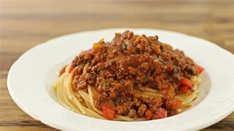 A white plate filled with spaghetti topped with a rich, chunky meat and tomato sauce. The sauce includes visible pieces of minced meat, diced tomatoes, and herbs, giving it a hearty appearance. The plate sits on a wooden table background.