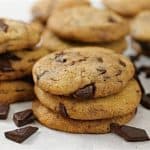 A close-up of stacks of chocolate chip cookies with chunks of chocolate scattered around them on a white surface. The cookies are golden-brown and look freshly baked. Multiple cookies are in the background, creating a visually appealing and appetizing display.