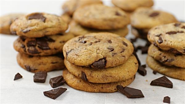 A close-up of stacks of chocolate chip cookies with chunks of chocolate scattered around them on a white surface. The cookies are golden-brown and look freshly baked. Multiple cookies are in the background, creating a visually appealing and appetizing display.