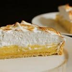Close-up of a slice of lemon meringue pie with a golden, slightly toasted meringue topping on a white plate. In the background, another slice of the pie is on a plate, slightly out of focus. The filling is a yellow lemon custard with a crust.