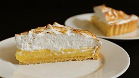 Close-up of a slice of lemon meringue pie with a golden, slightly toasted meringue topping on a white plate. In the background, another slice of the pie is on a plate, slightly out of focus. The filling is a yellow lemon custard with a crust.