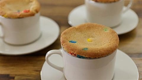 Three white cups with matching saucers containing cookies with colorful candy pieces on top. The cookies appear to be freshly baked, resting on the edges of the cups. The scene is set on a wooden table.