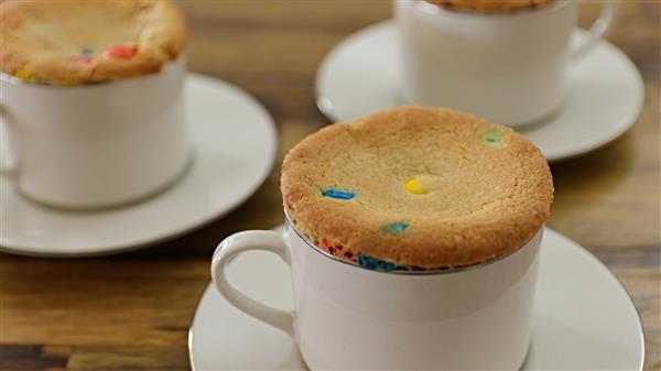 Three white cups with matching saucers containing cookies with colorful candy pieces on top. The cookies appear to be freshly baked, resting on the edges of the cups. The scene is set on a wooden table.