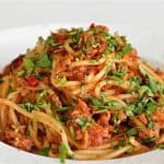 A plate of spaghetti topped with a savory sauce, shredded chicken, and garnished with fresh chopped parsley and red chili flakes. The dish is presented on a white plate against a plain background.