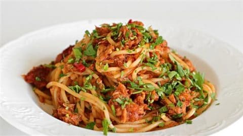 A plate of spaghetti topped with a savory sauce, shredded chicken, and garnished with fresh chopped parsley and red chili flakes. The dish is presented on a white plate against a plain background.