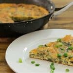 A slice of vegetable frittata garnished with fresh green onions sits on a white plate. Behind it, the rest of the frittata is in a black skillet on a wooden surface. The frittata appears to have spinach and mushrooms among other ingredients.