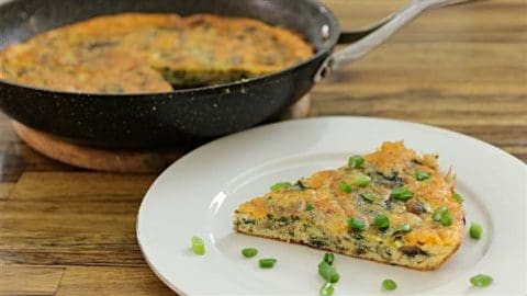 A slice of vegetable frittata garnished with fresh green onions sits on a white plate. Behind it, the rest of the frittata is in a black skillet on a wooden surface. The frittata appears to have spinach and mushrooms among other ingredients.