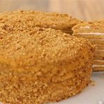 A layered honey cake with a golden crumb exterior sits on a white plate. A slice has been cut from the cake, revealing multiple layers with cream filling between them. The cake is presented on a wooden surface.