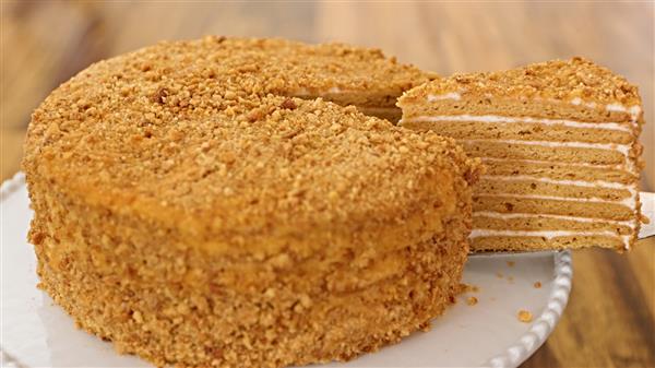 A layered honey cake with a golden crumb exterior sits on a white plate. A slice has been cut from the cake, revealing multiple layers with cream filling between them. The cake is presented on a wooden surface.