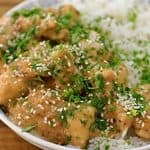 A bowl of rice topped with pieces of cooked chicken covered in a creamy sauce, garnished with chopped green herbs and sesame seeds. The dish is set on a wooden table.