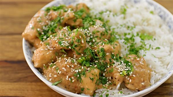 A bowl of rice topped with pieces of cooked chicken covered in a creamy sauce, garnished with chopped green herbs and sesame seeds. The dish is set on a wooden table.