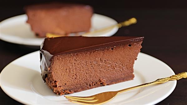 A slice of rich chocolate cheesecake with a smooth, glossy chocolate topping sits on a white plate with a gold fork beside it. Another identical slice is blurred in the background on a similar plate. The background is dark, highlighting the dessert.