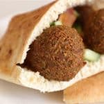 A close-up of a pita bread filled with two crispy falafel balls, chopped cucumber, and tomato, placed on a white plate. The falafel and vegetables peek out from the fold of the pita, showcasing a fresh and appetizing Mediterranean dish.