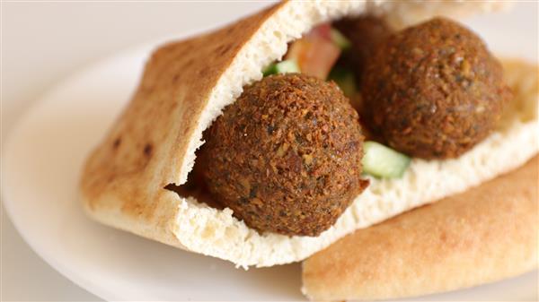 A close-up of a pita bread filled with two crispy falafel balls, chopped cucumber, and tomato, placed on a white plate. The falafel and vegetables peek out from the fold of the pita, showcasing a fresh and appetizing Mediterranean dish.