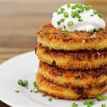 Four golden-brown, crispy potato cakes stacked on a white plate, topped with a dollop of sour cream and garnished with chopped chives, sitting on a wooden table.