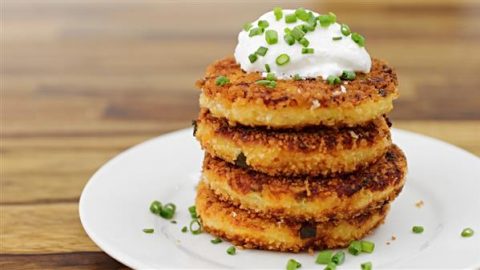 Four golden-brown, crispy potato cakes stacked on a white plate, topped with a dollop of sour cream and garnished with chopped chives, sitting on a wooden table.