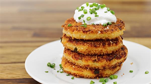 Four golden-brown, crispy potato cakes stacked on a white plate, topped with a dollop of sour cream and garnished with chopped chives, sitting on a wooden table.