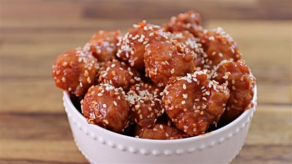 A white bowl filled with golden-brown, bite-sized pieces of food covered in a glossy red sauce and sprinkled with sesame seeds. The bowl is placed on a wooden surface.