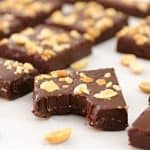 Several rectangular chocolate brownies topped with chopped peanuts are arranged on a light-colored surface. One brownie in the foreground has a bite taken out of it. Scattered peanuts are visible among the brownies.