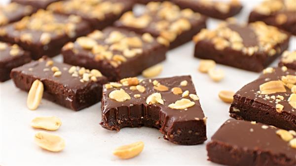 Several rectangular chocolate brownies topped with chopped peanuts are arranged on a light-colored surface. One brownie in the foreground has a bite taken out of it. Scattered peanuts are visible among the brownies.