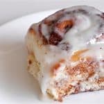 A cinnamon roll with white icing sits on a white plate. The roll is soft and fluffy, with visible layers of cinnamon filling swirled throughout. The icing is generously drizzled on top, partially melting and adding a glossy, sweet finish.