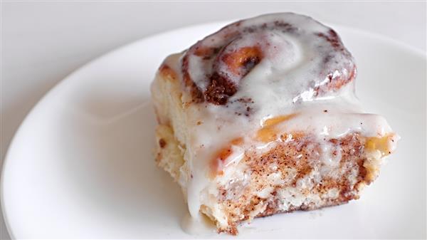 A cinnamon roll with white icing sits on a white plate. The roll is soft and fluffy, with visible layers of cinnamon filling swirled throughout. The icing is generously drizzled on top, partially melting and adding a glossy, sweet finish.