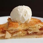 A slice of apple pie topped with a scoop of vanilla ice cream on a white plate. The pie has a golden-brown crust and visible slices of apple with a cinnamon filling. The background is dark, making the dessert stand out.