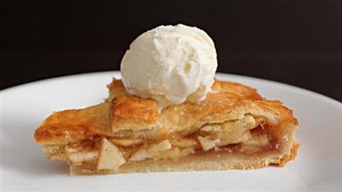 A slice of apple pie topped with a scoop of vanilla ice cream on a white plate. The pie has a golden-brown crust and visible slices of apple with a cinnamon filling. The background is dark, making the dessert stand out.