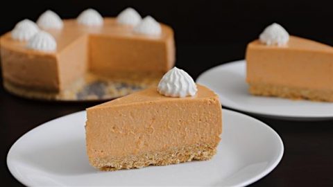 A close-up of a sliced pumpkin cheesecake with a graham cracker crust. The main slice is on a white plate in the foreground, topped with a dollop of whipped cream. The rest of the cheesecake and another slice are visible in the background.