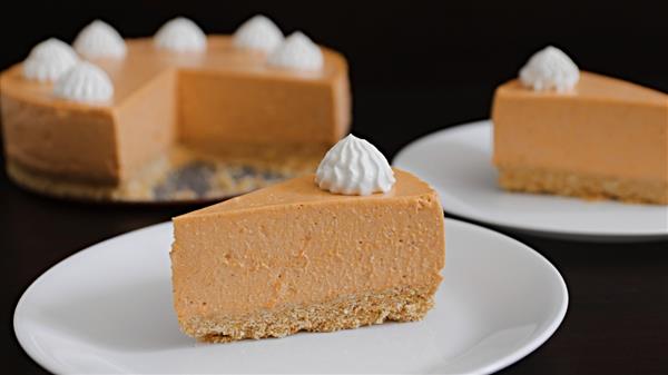 A close-up of a sliced pumpkin cheesecake with a graham cracker crust. The main slice is on a white plate in the foreground, topped with a dollop of whipped cream. The rest of the cheesecake and another slice are visible in the background.