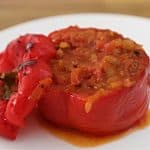A stuffed red bell pepper is presented on a white plate. The pepper is filled with a savory mixture of rice, onions, and tomato sauce. To the side, a roasted cap of the bell pepper is placed, completing the dish. The wooden table is visible in the background.