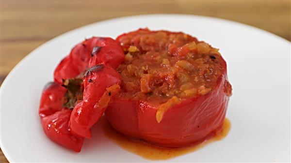 A stuffed red bell pepper is presented on a white plate. The pepper is filled with a savory mixture of rice, onions, and tomato sauce. To the side, a roasted cap of the bell pepper is placed, completing the dish. The wooden table is visible in the background.